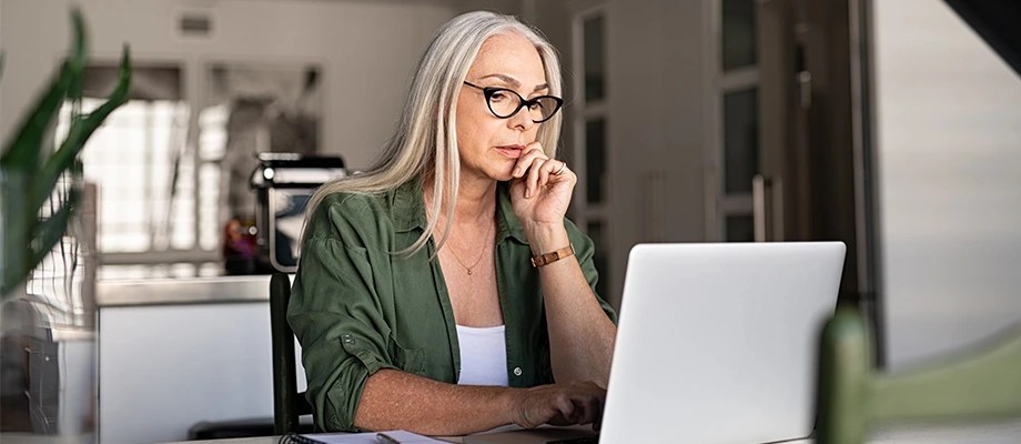 Femeie cu par alb si ochelari lucrand concentrata la laptop intr-un spatiu modern
